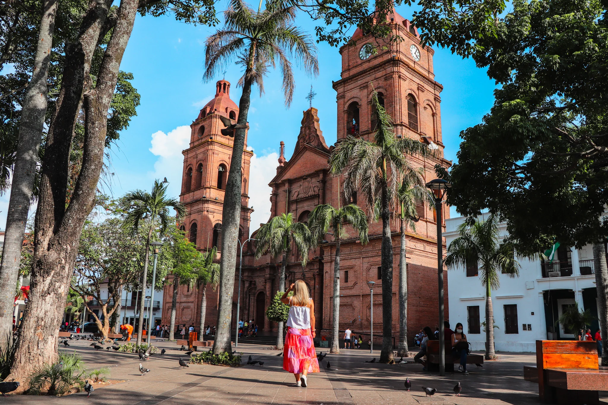 A blonde woman walking away from the camera in a plaza towards a large brick cathedral.